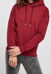 Sudadera roja con capucha, cordones con puntas doradas. Tela suave, ajuste relajado, puños y dobladillo acanalados, combinada con jeans grises.