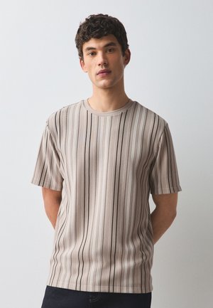 Ung mand med krøllet hår iført en beige og hvid kortærmet skjorte med lodrette striber, stående mod en ensfarvet lys baggrund.