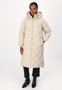 Beige gewatteerde lange jas met een capuchon, voorzien van een rits aan de voorkant, elastische manchetten en zijzakken. Textuurstof en diamanten patroon details.