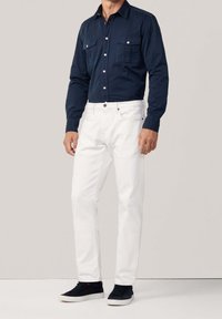 Marineblaues Langarmhemd mit Knöpfen und zwei Brusttaschen, kombiniert mit weißen Jeans und schwarzen Sneakers. Das Hemd hat einen Kragen und eine figurbetonte Passform.