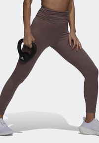 Brune højtaljede leggings med rynkning, lavet af elastisk materiale; holder en sort kettlebell. Hvide sportssko synlige.