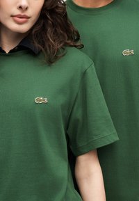 Δύο άτομα που φορούν πανομοιότυπα πράσινα t-shirt Lacoste, με το κεντημένο λογότυπο κροκοδείλου στο αριστερό στήθος.