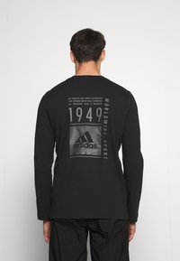 Musta pitkähihainen paita, jossa on graafinen design selässä, mukaan lukien teksti ja Adidas-logot, sekä sileä pinta.