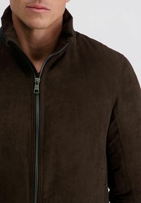 Braune Wildlederjacke mit hohem Kragen, vorderem Reißverschluss und glatter Textur. Entworfen mit dezenten Nähdetails und ohne sichtbare Logos.