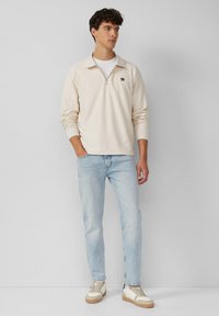 Hellbeiger Pullover mit Reißverschlusskragen, strukturiertem Stoff, kombiniert mit hellblauen Jeans und beigen Sneakers mit braunen Akzenten.