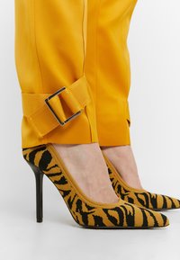 Chaussures à talons hauts avec imprimé animal noir et orange, portées avec un pantalon orange vif doté de sangles réglables à boucle aux chevilles.