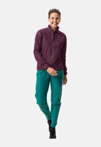 Une personne souriante marchant, portant une veste zippée violette, un pantalon teal et des chaussures noires sur un fond léger uni.