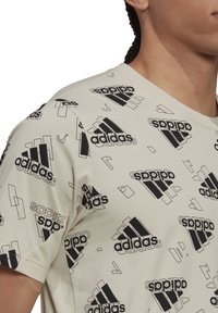 Kaksi vaaleanbeige T-paitaa, joissa on mustaa adidas-logoa ja abstrakteja geometrisia muotoja kauttaaltaan. Valmistettu sileästä puuvillakankaasta, pyöreä pääntie.