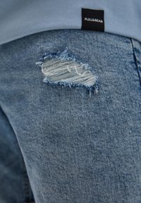 Gros plan sur un jean en denim bleu clair avec un détail de déchirure effiloché près de la cuisse et une chemise bleue avec une étiquette Pull&Bear.