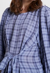 Blåternet bluse med bånd i taljen og flagrende ærmer. Stoffet har en blød tekstur med en blanding af lyse og mørke blå firkanter.