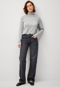 Maglione grigio a collo alto, abbinato a jeans a gamba larga grigi scuri. Modella in piedi con le mani sui fianchi, indossando ballerine nere. Sfondo neutro.