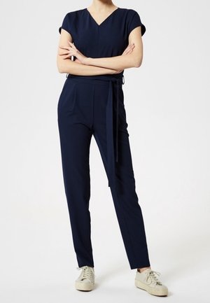 Personne portant une combinaison à manches courtes bleu marine avec ceinture à la taille, debout les bras croisés et des baskets blanches sur un fond blanc.