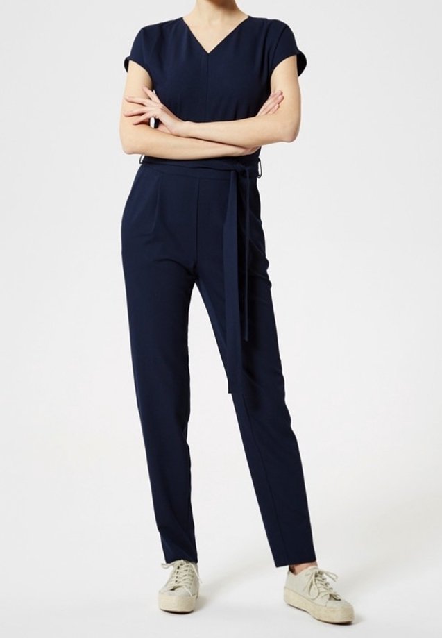 Personne portant une combinaison à manches courtes bleu marine avec ceinture à la taille, debout les bras croisés et des baskets blanches sur un fond blanc.