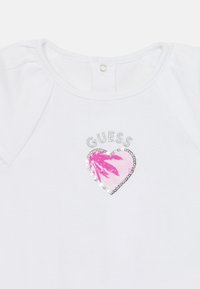 Weißes T-Shirt mit kurzen Ärmeln, das ein pinkes Herzmotiv mit Palmblattdruck zeigt, umgeben von silbernen Pailletten, und einen Druckverschluss am Hals hat.