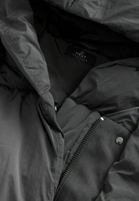Tamsiai pilka lietaus striukė su lygiu paviršiumi, turinti zip užtrauktuką, prispaudimo aparatūrą ir etiketę su užrašu „Next Rainwear Collection“.