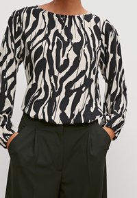 Blouse à manches longues à motif noir et blanc avec un col rond et des détails froncés, associée à un pantalon taille haute vert foncé.
