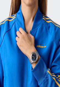 Sudraba rokas pulkstenis ar zilu ciparu, nerūsējošā tērauda sietiņa siksna, kas apvienota ar spilgtu zilu jaku ar rāvējslēdzēju, kurai ir dzelteni svīteri un logo.