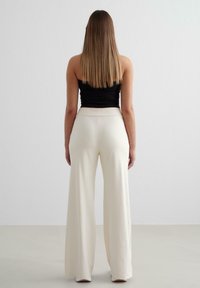 Witte wijde broek van een zachte, gladde stof, met een hoge tailleband. Het model draagt een aansluitend zwart topje en staat met de rug naar de camera.