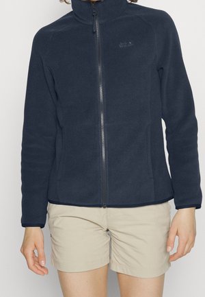 Persoon die een marineblauwe fleece jas met ritssluiting en beige korte broek draagt, zichtbaar vanaf de nek tot halverwege de dij, tegen een effen achtergrond.