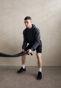 Felpa grigia con cappuccio e pantaloni corti neri, con corde da allenamento nere. Modello con scarpe nere e calzini bianchi, in piedi su un pavimento liscio.