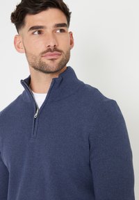 Dunkelblauer Pullover mit Reißverschlusskragen aus weichem, strukturiertem Strick. Mit einem Stehkragen und einem kleinen silbernen Reißverschlussdetail.