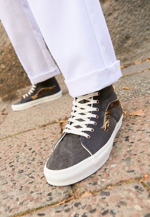 Baskets montantes en toile gris foncé avec des accents à imprimé animalier, des lacets blancs et une semelle en caoutchouc texturée. Portées avec un pantalon blanc retroussé.