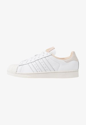 Biały niski sneaker z ochronnym noskiem, trzema przeszywanymi paskami, zapięciem na sznurowadła oraz beżowymi akcentami na pięcie i języku.