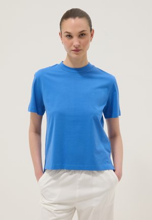 Jeune personne aux cheveux attachés porte une chemise bleue à manches courtes et un pantalon blanc, debout les mains dans les poches sur un fond uni.