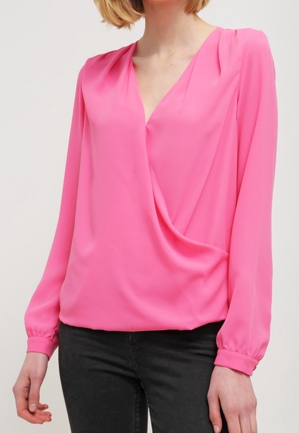 Femme portant un chemisier rose vif à manches longues avec un col en V et un devant drapé, associé à un pantalon noir sur un fond uni.