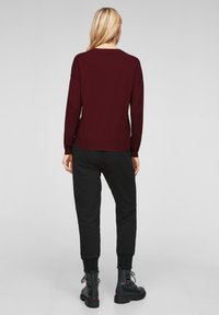 Frau mit blondem Haar, die einen burgunderfarbenen Pullover, schwarze Hosen und schwarze Kampfstiefel trägt, steht mit dem Rücken zur Kamera vor einem grauen Hintergrund.