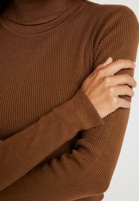 Pull en maille côtelée marron à col cheminée avec des manches longues, présentant des poignets ajustés et une finition texturée, mettant en avant des lignes verticales et un matériau doux.