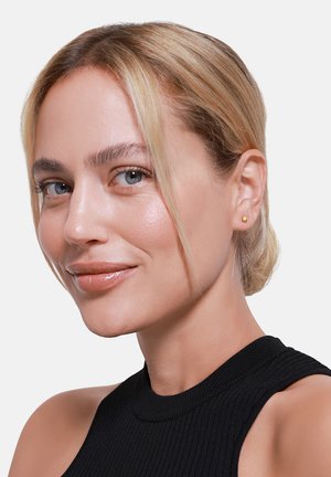 Goldstudohrring, der am Ohr eines Models positioniert ist, mit einer einfachen runden Form und glatter Oberfläche. Der Hintergrund ist rein weiß, was den Ohrschmuck betont.