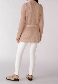 Cardigan de punto rosa claro con cinturón, mangas largas y textura acanalada, combinado con pantalones blancos de corte ajustado y sandalias beige.