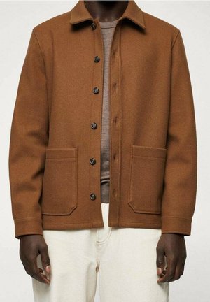 Mężczyzna ubrany w brązową wełnianą kurtkę zapinaną na guziki z dwoma przednimi kieszeniami, pod którą ma beżową koszulę, oraz w jasnobeżowe spodnie.