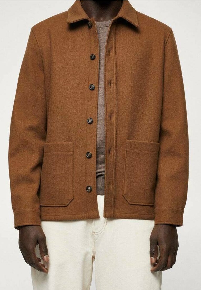 Homme portant une veste en laine marron à boutons avec deux poches avant, une chemise beige en dessous et un pantalon blanc cassé.