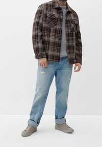 Mann in einem braun-gestreiften Jackett mit grauem Sweater, hellblauen, hochgekrempelten Jeans mit Löchern und beigen Turnschuhen, der vor einem weißen Hintergrund steht.