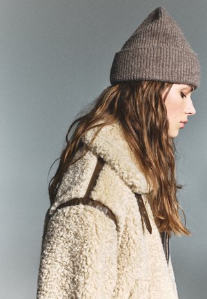Beige shearling-takk med brune skinnakenter, en ribbet brun strikket lue, og langt bølgete hår synlig mot en grå bakgrunn.