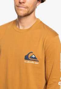 Long-sleeve mustard shirt made of soft fabric, featuring a blue graphic logo and text on the left sleeve. Simple, fitted design.