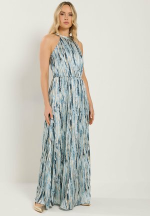 Vrouw draagt een mouwloze, vloerlange halterjurk met blauwe en beige patronen, staande tegen een effen lichte achtergrond.