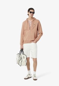Sudadera rosa claro con cremallera y bolsillos frontales, combinada con una camiseta de estampado, pantalones cortos blancos y un bolso crema con acentos negros.
