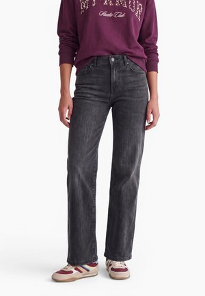 Personne portant un jean droit gris foncé, un sweat bordeaux avec un motif léopard, et des baskets blanches avec des rayures rouges et bleues.