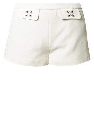 Femme marchant portant un haut noir, un short blanc avec des rabats décoratifs, des collants noirs, des bottines noires vernies et un sac à bandoulière noir clouté.