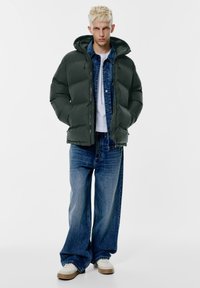 Donkergroene gewatteerde jas met capuchon, gedragen over een denim overhemd, gecombineerd met losse blauwe jeans en witte sneakers tegen een effen achtergrond.