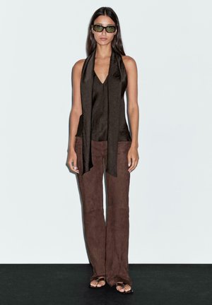 Brown silk top with a v-neck and long scarf, paired with brown suede wide-leg pants and flat sandals. Model stands against a neutral background.