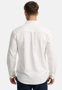 Camisa de lino blanca con mangas largas, puños abotonados y un pliegue en la parte posterior. La tela tiene una ligera textura y un ajuste relajado.