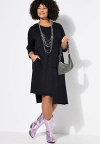 Vestido negro casual con mangas de tres cuartos, bolsillos y un dobladillo trasero más largo. Combinado con un collar plateado en capas y botas metálicas rosas.