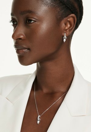 Primer plano de una mujer que lleva un blazer blanco, pendientes colgantes de diamantes y un collar a juego con colgantes en forma de lágrima.