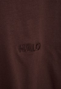 Bruine katoenstof met een gladde textuur, voorzien van een geborduurd "HALO"-logo in een iets donkerdere tint. Eenvoudig en minimalistisch ontwerp.