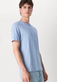 T-shirt de mangas curtas azul claro, em um tecido macio e texturizado. Apresenta um decote redondo e um pequeno logotipo no peito. Corte casual.