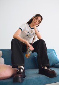 White T-shirt with navy trim featuring cartoon characters, paired with oversized dark denim pants and chunky black platform shoes. Clear drink cup in hand.